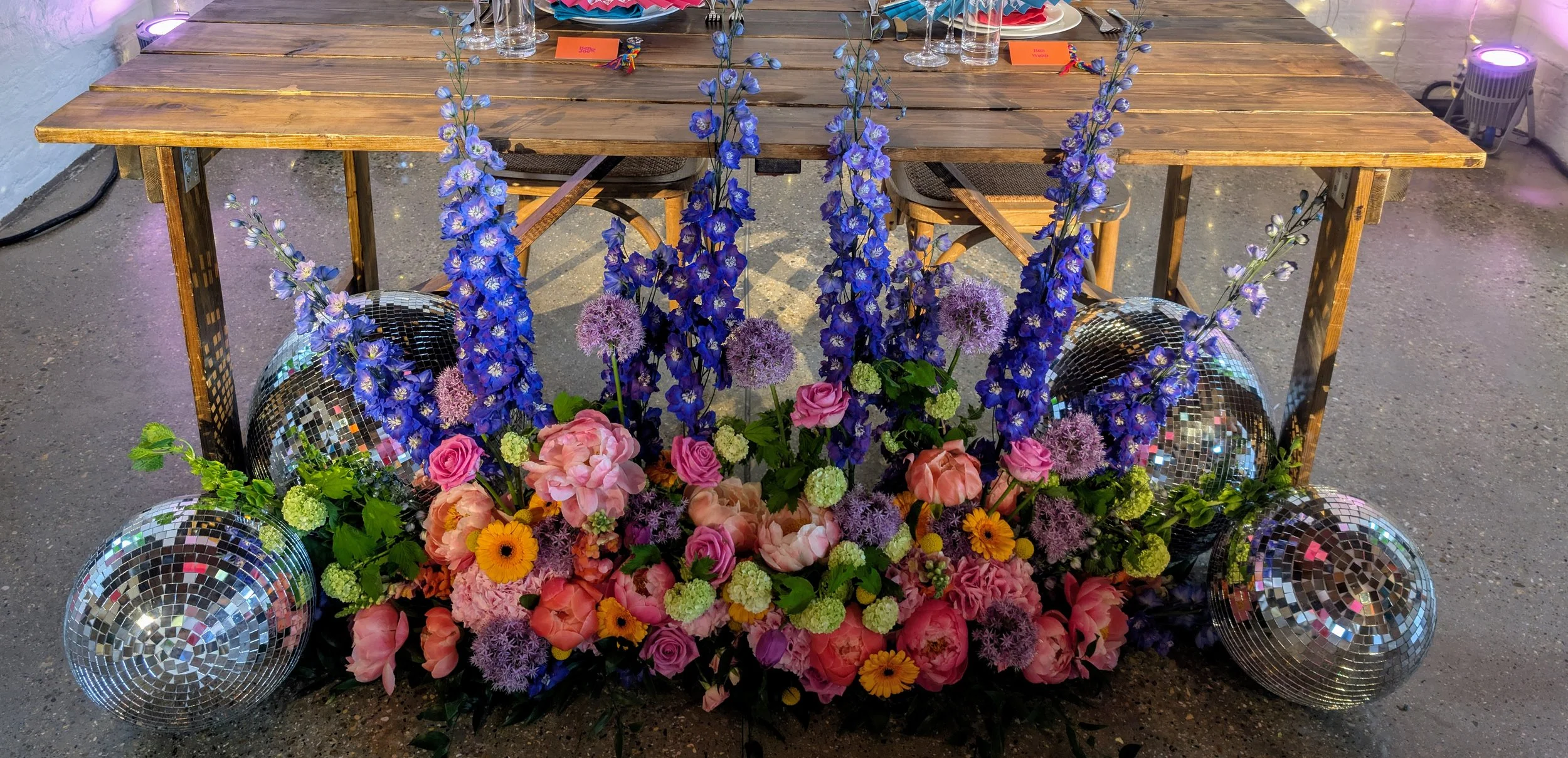 Colourful tablescape floral arrangement by Harlequin Florals featuring blue delphiniums, pink peonies, purple alliums, and orange gerberas accented with disco balls. Styled beneath a rustic wooden table, blending bold blooms with retro party vibes fo