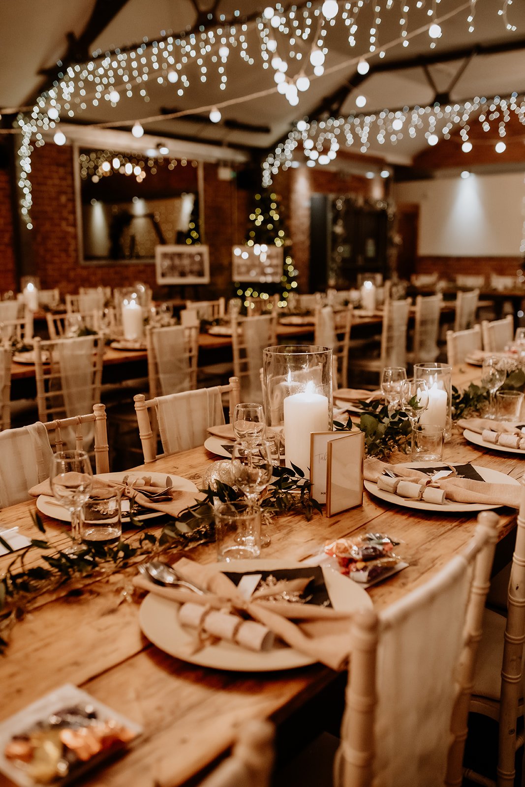 Long wooden tables are set for dinner with neutral plates, soft napkins and small crackers at each place, while leafy green garlands and tall glass candles run down the centre and warm fairy lights drape overhead, creating a cosy golden glow across t