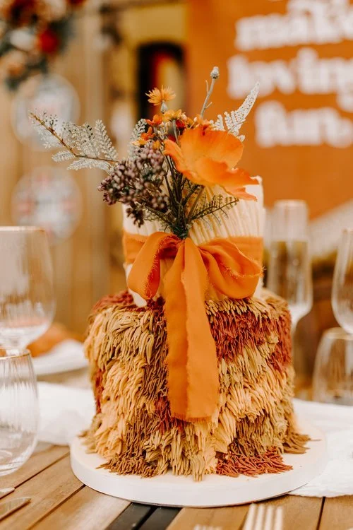 A bold and textured creation by Amber’s Cakes and Treats, this two-tier masterpiece bursts with autumnal warmth and bohemian flair. The bottom tier features cascading shag-style buttercream piping in rich tones of burnt orange, caramel, and cream, wh