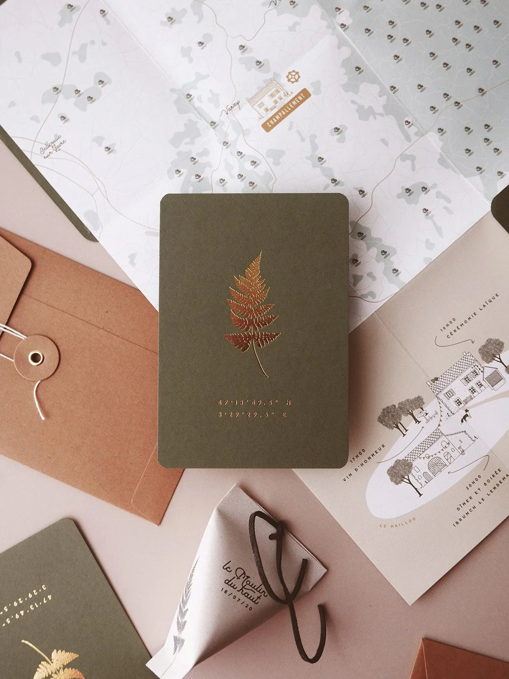 A beautifully styled wedding invitation suite featuring an olive green card with a gold foiled fern motif surrounded by illustrated venue maps, kraft envelopes and a personalised confetti cone creating a refined nature inspired aesthetic perfect for 