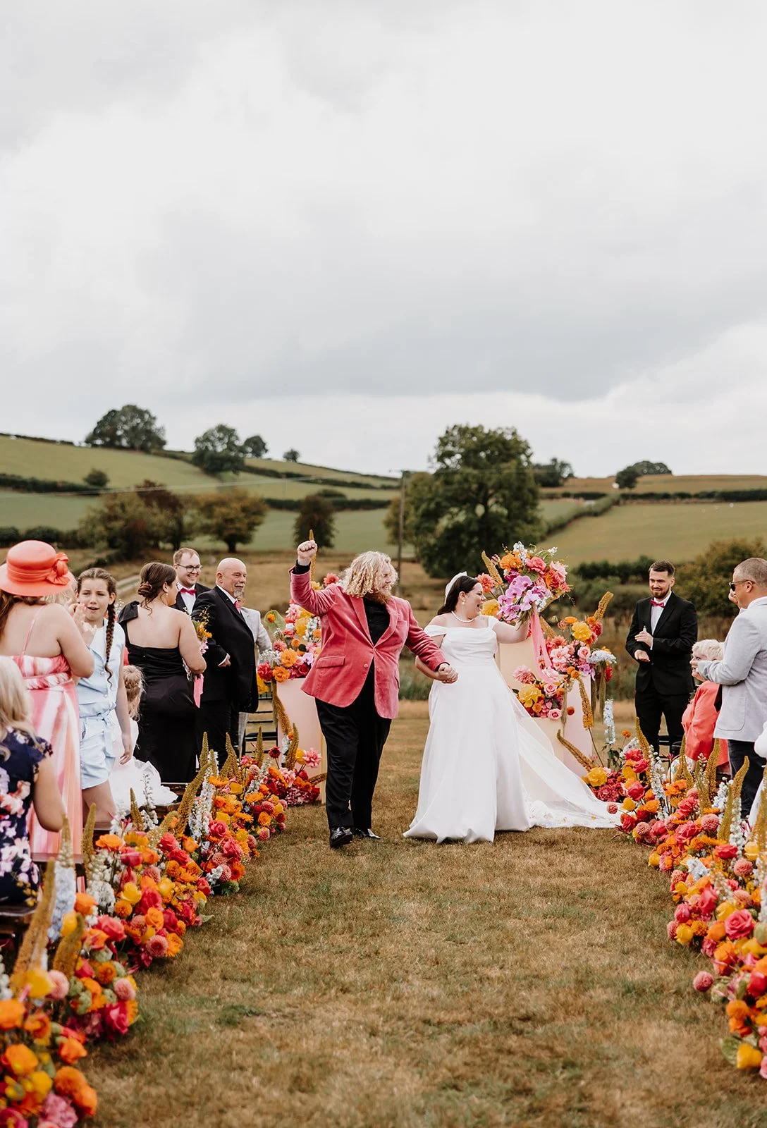 A joyful outdoor wedding ceremony at The Giraffe Shed in Wales, featuring a newlywed couple walking down the aisle surrounded by vibrant floral arrangements in pink, orange, and yellow hues. The lush countryside backdrop and creative aisle decor set 