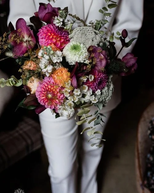 Vibrant modern wedding bouquet by New Bud Floristry featuring bold dahlias, hellebores, queen anne’s lace, roses and textured foliage in rich pink, purple and peach tones. Shown against a crisp white suit, this striking garden inspired arrangement hi