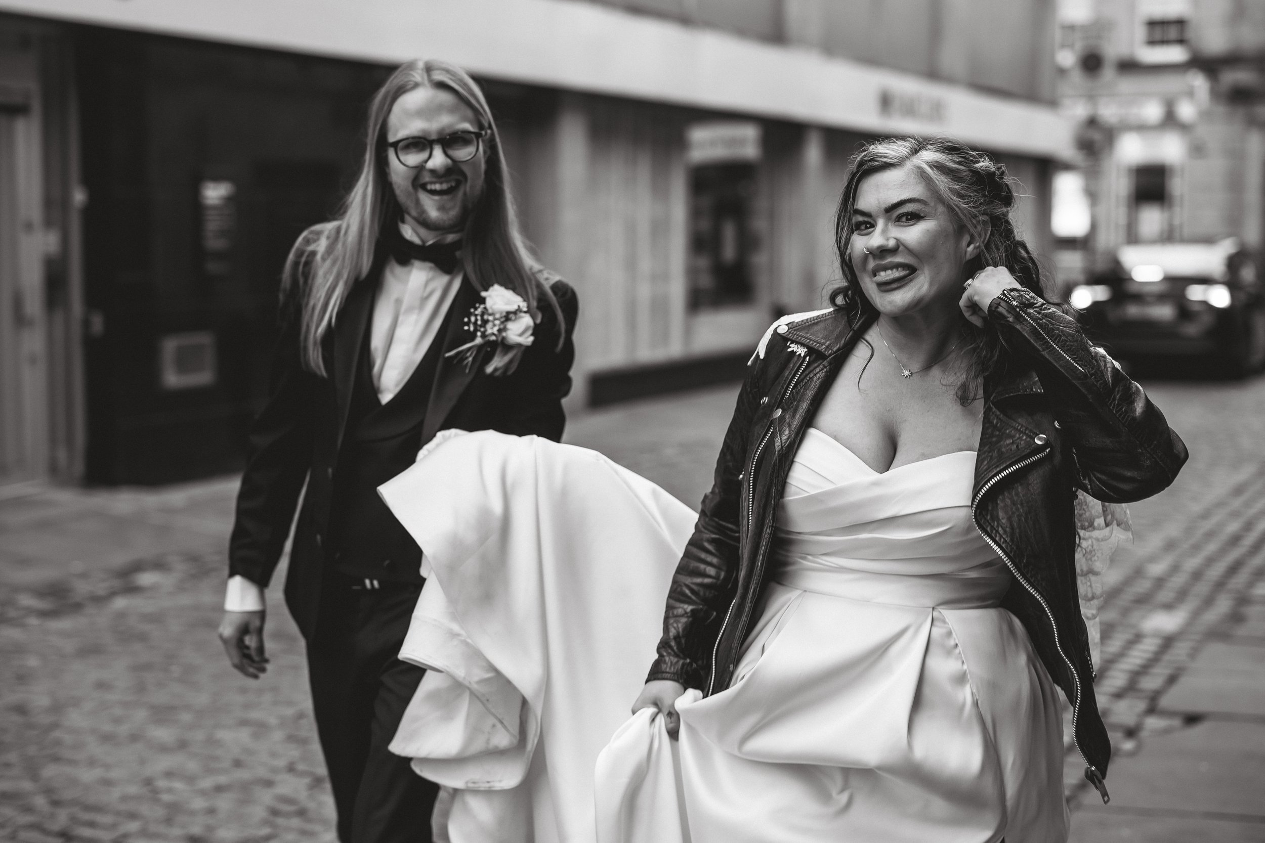 A candid and stylish wedding moment captured by Joy Louise Photography, showing a newly married couple walking through the city streets with relaxed confidence. The bride wears a flowing white dress paired with a black leather jacket, smiling broadly