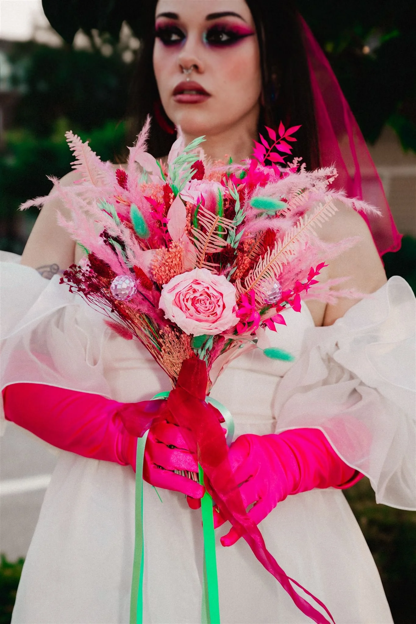A striking and dramatic bridal portrait featuring a vivid bouquet in bold hot pink, red, mint and soft blush tones, filled with feathery textures, preserved foliage, playful dyed stems and a single central rose that anchors the arrangement, with tiny