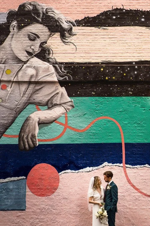 A couple stands together beneath a huge, vibrant mural that stretches across the brick wall behind them. The artwork shows a large figure painted in soft tones, with flowing hair and an outstretched arm that seems to hover protectively above the pair