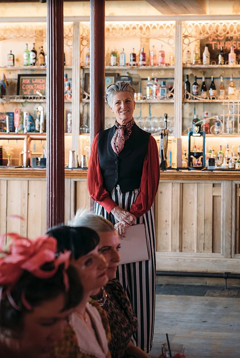 Arabella stands near the bar holding her notes, dressed in a waistcoat and striped trousers, watching the room with a calm, attentive expression as guests sit in the foreground. Joanna Bongard Photography.