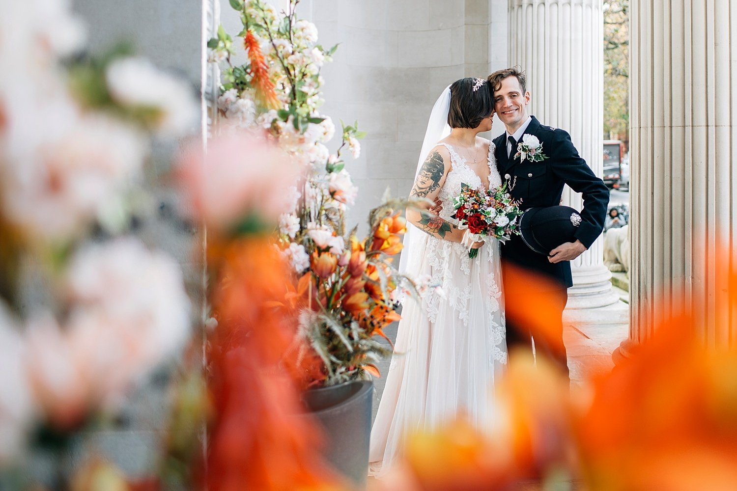 A burst of soft, cinematic colour frames this intimate moment as the couple stands between grand stone columns, wrapped in petals and bright florals. They’re tucked in close, sharing a quiet smile that says everything - her lace gown and bold tattoos