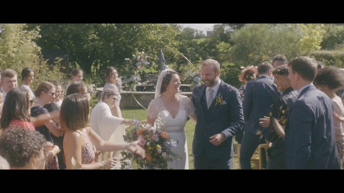 A newly married couple walks through a sunlit garden surrounded by smiling guests who toss handfuls of confetti while soft florals vibrant greenery and warm summer light blend into a gentle cinematic scene filled with easy laughter and heartfelt cele