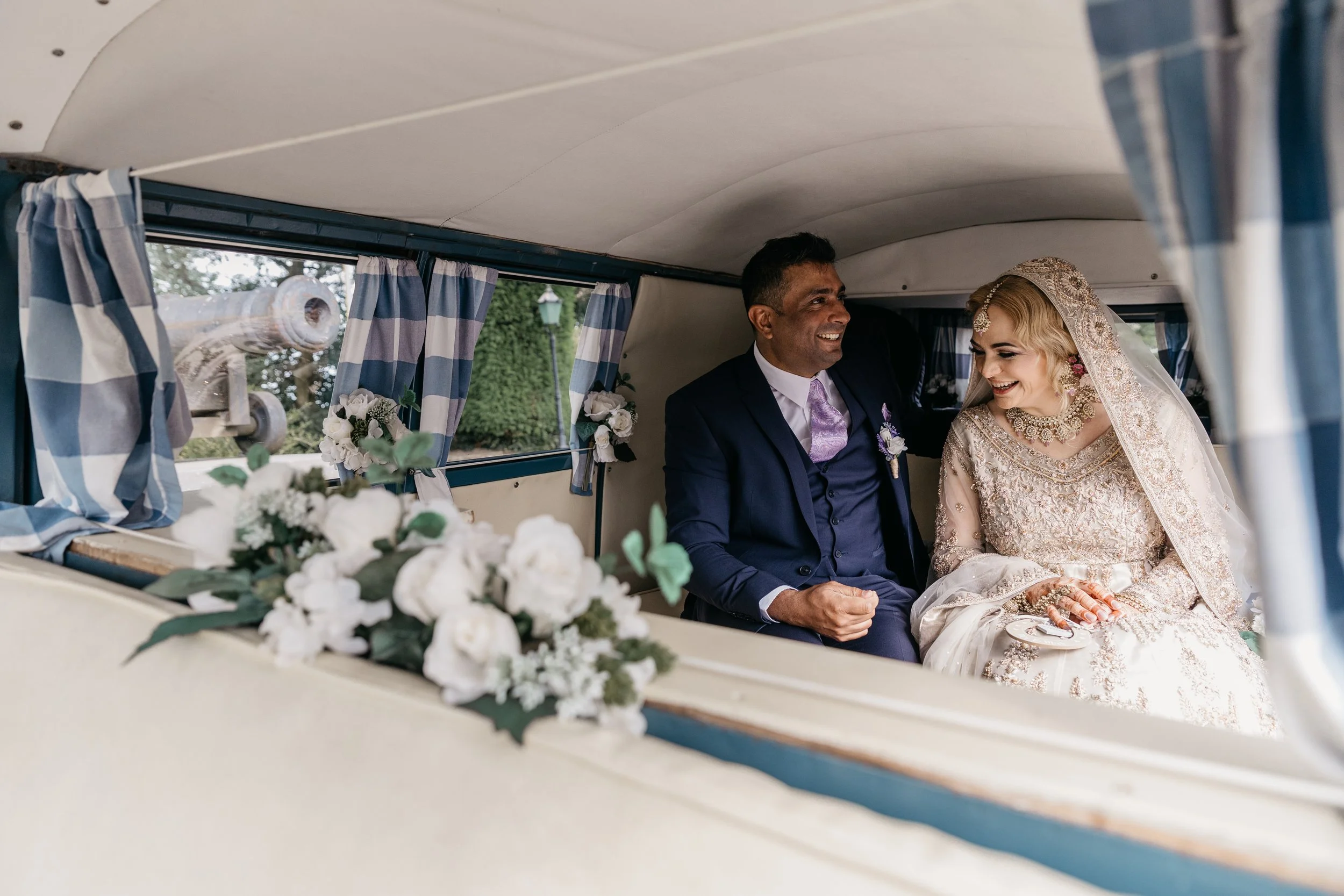 A tender and joy filled moment captured by YU Photography, showing the couple sharing a warm smile together inside a beautifully decorated vintage vehicle. Soft blue checked curtains frame the windows while white florals add a romantic touch to the c