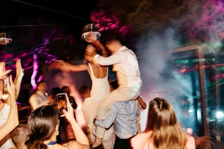 Guests lifting Mr and Mrs L onto shoulders during an outdoor wedding party, surrounded by colourful lighting, smoke effects, and disco balls. The energetic scene captures the excitement of a packed dance floor, perfect for couples wanting unforgettab