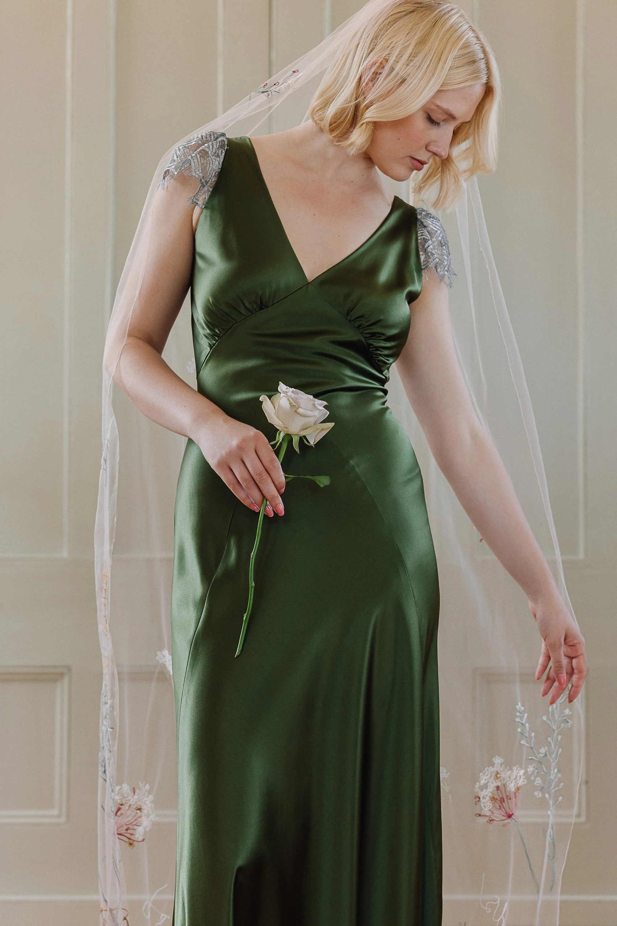 The model wears a deep green bias cut silk gown that drapes smoothly over her figure, its soft sheen catching the light while delicate silver lace accents sit on the shoulders, and she holds a single pale rose as a long embroidered veil falls around 