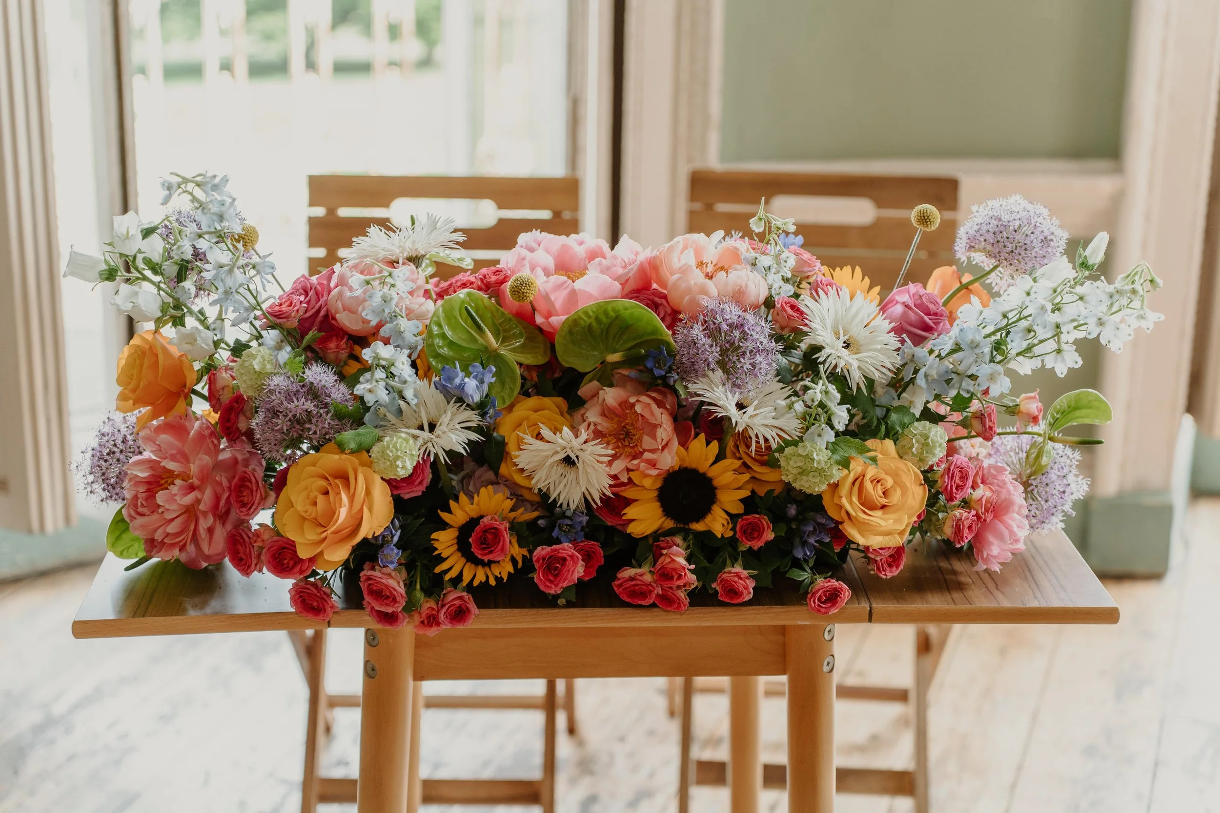 Colourful wedding ceremony arrangement by Harlequin Florals, photographed by Alina Pullen Photography. A mix of sunflowers, roses, peonies, delphiniums, and anthuriums in bright summer shades of yellow, pink, orange, and blue, adorning a rustic woode