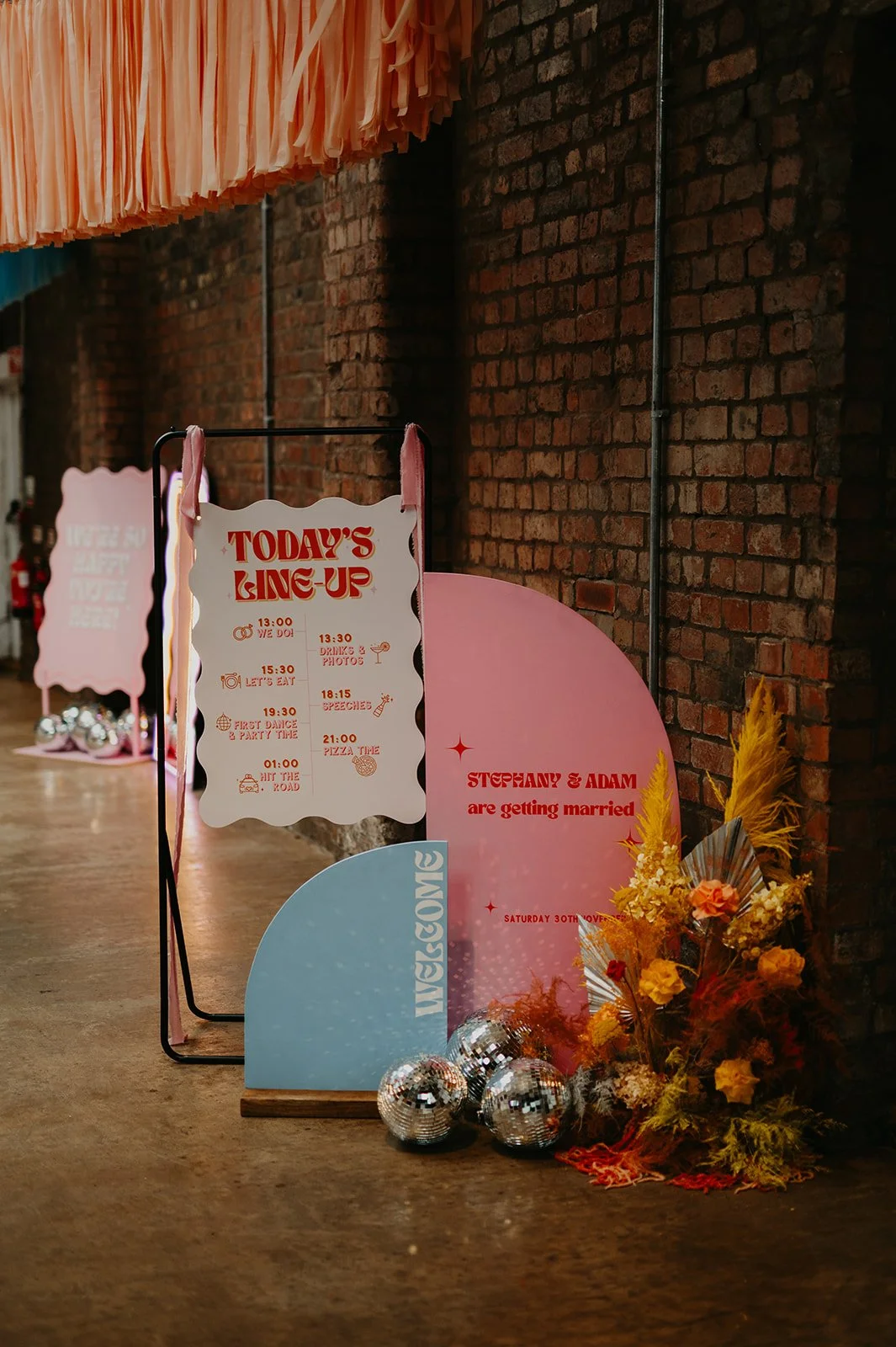 Vibrant wedding signage display by AMW Events featuring a retro-style pink and white schedule board, colourful welcome signs in pink and blue, disco balls, and a bold yellow-orange floral arrangement against an exposed brick wall.