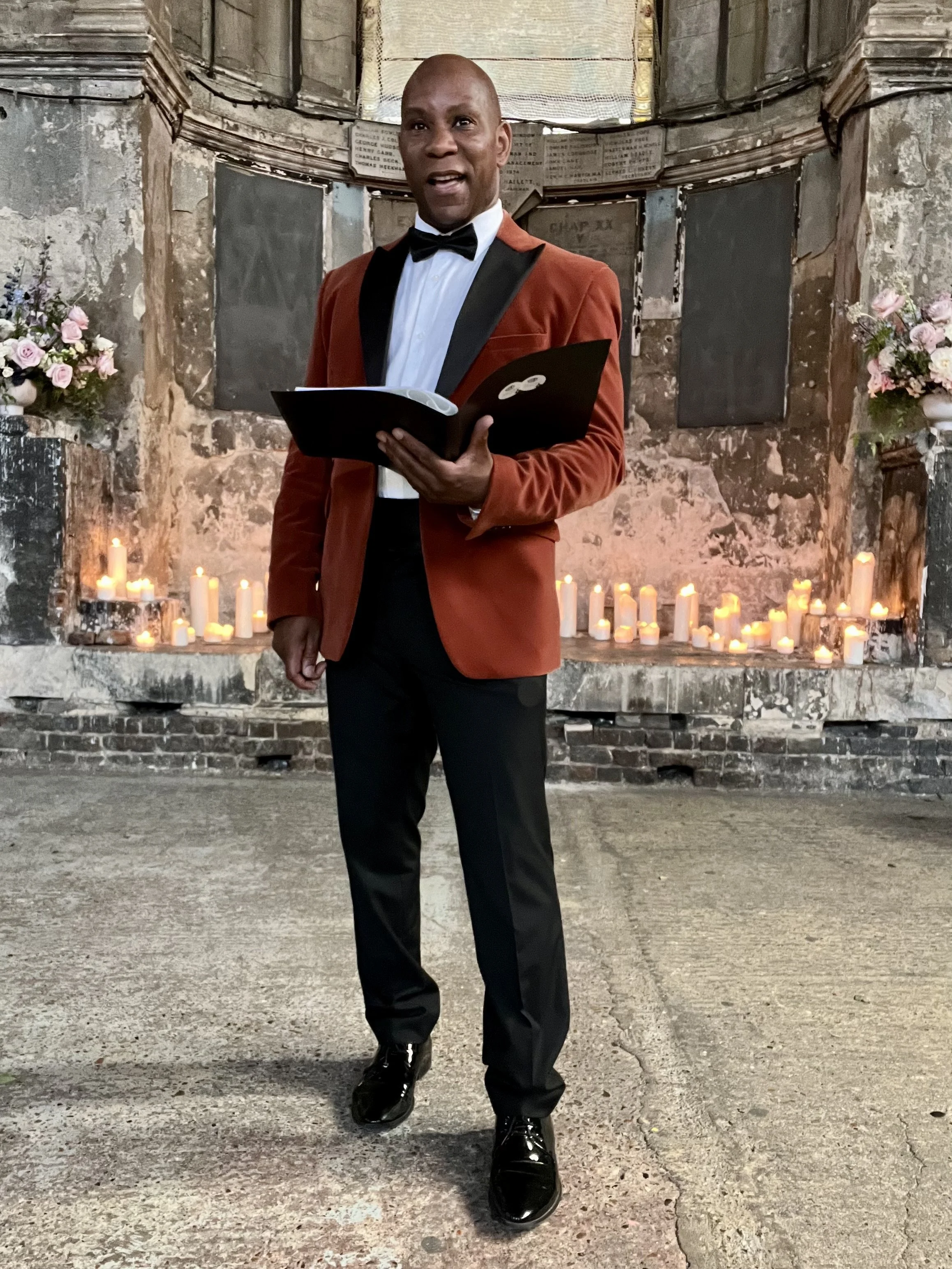 Dave Octave Celebrant in a red blazer, standing in front of a candle lit altar, reading from an order of service. 