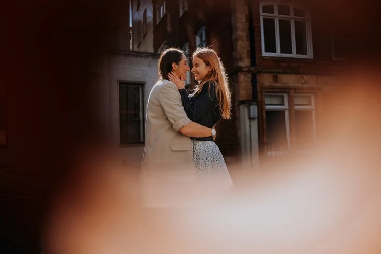 A couple embrace in a quiet city street with warm soft foreground framing, captured by Joshua Humphrey Photography. Their smiles and gentle connection create a romantic candid portrait with dreamy light and an intimate urban atmosphere.