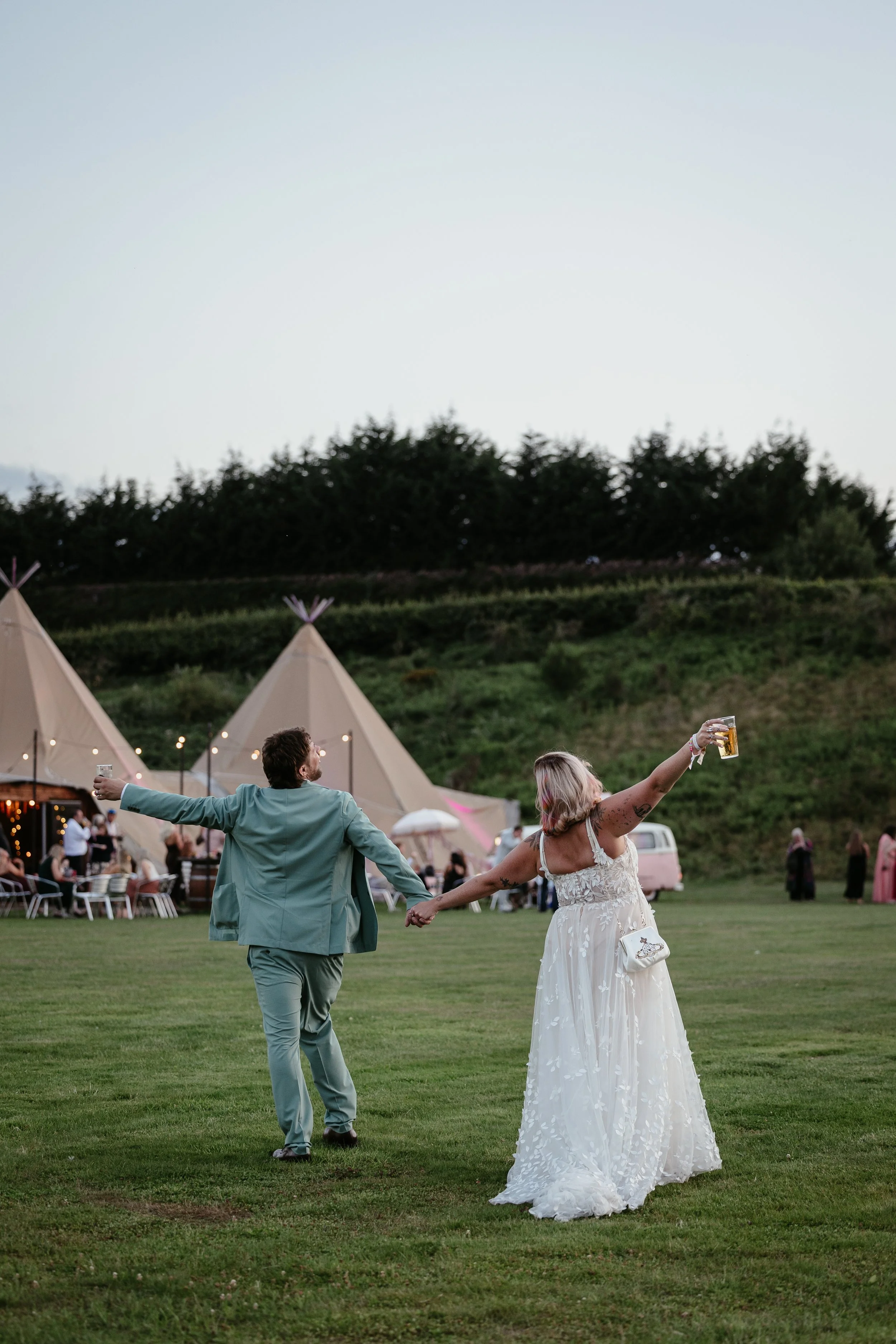 A joyful and carefree moment captured by Joy Louise Photography, showing the newly married couple walking hand in hand across an open field at their outdoor tipi wedding. With their backs to the camera, they lift their arms in celebration, each holdi