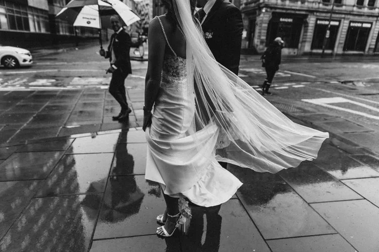 A cinematic and atmospheric street moment captured by YU Photography, showing the bride and groom standing on rain soaked pavements with the bride’s veil sweeping dramatically in the wind. Reflections shimmer across the wet ground as passersby move t