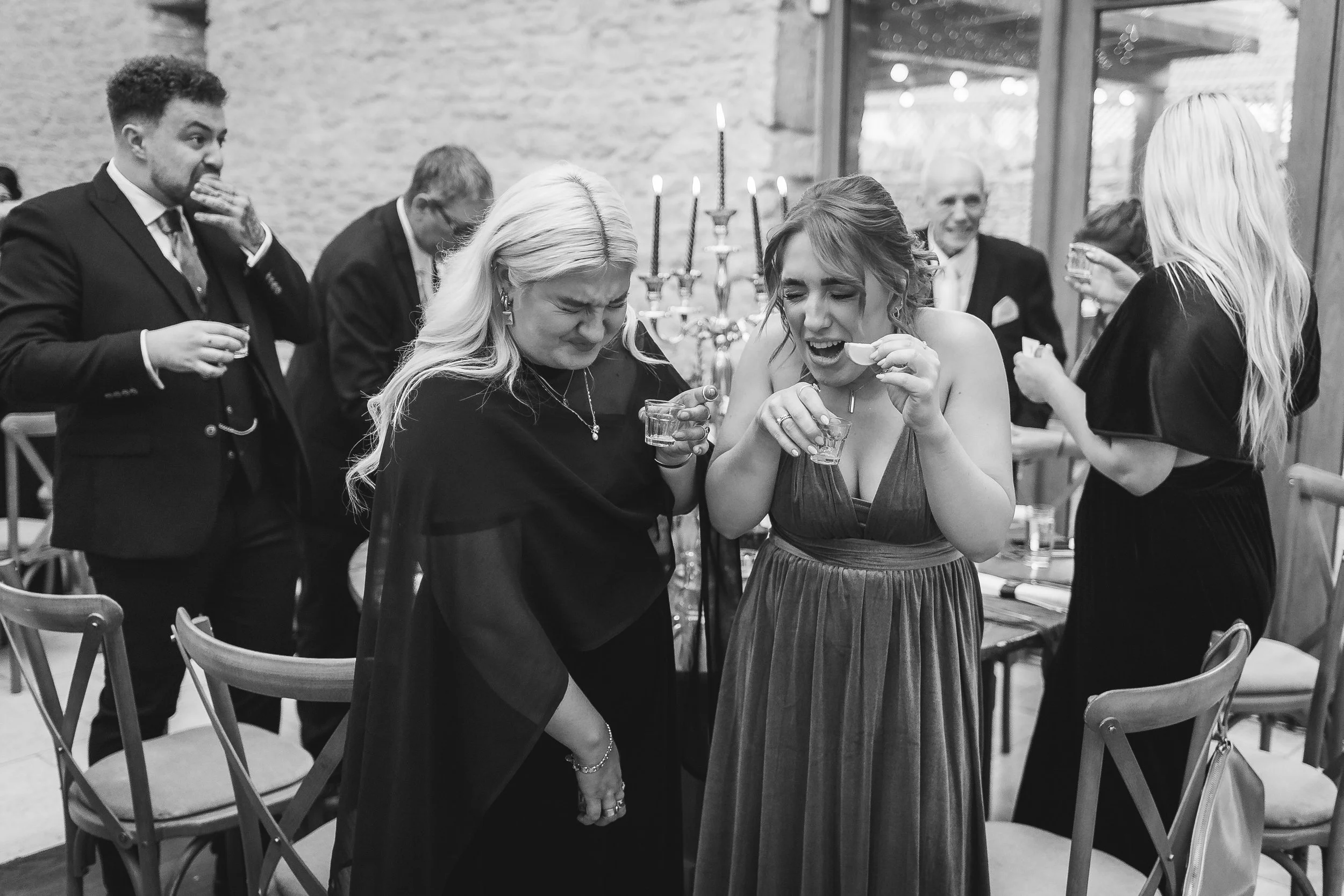 A candid black-and-white wedding reception moment captured by Gemma Gaskins Photography, featuring guests laughing and reacting dramatically while taking celebratory shots. The natural, documentary-style storytelling shows genuine emotion, fun, and t
