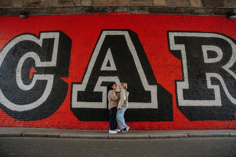 A couple stand close together in front of a bold red and black street-art mural under a railway arch, captured by Joshua Humphrey Photography. The graphic urban backdrop and their relaxed connection create a striking modern portrait perfect for alter