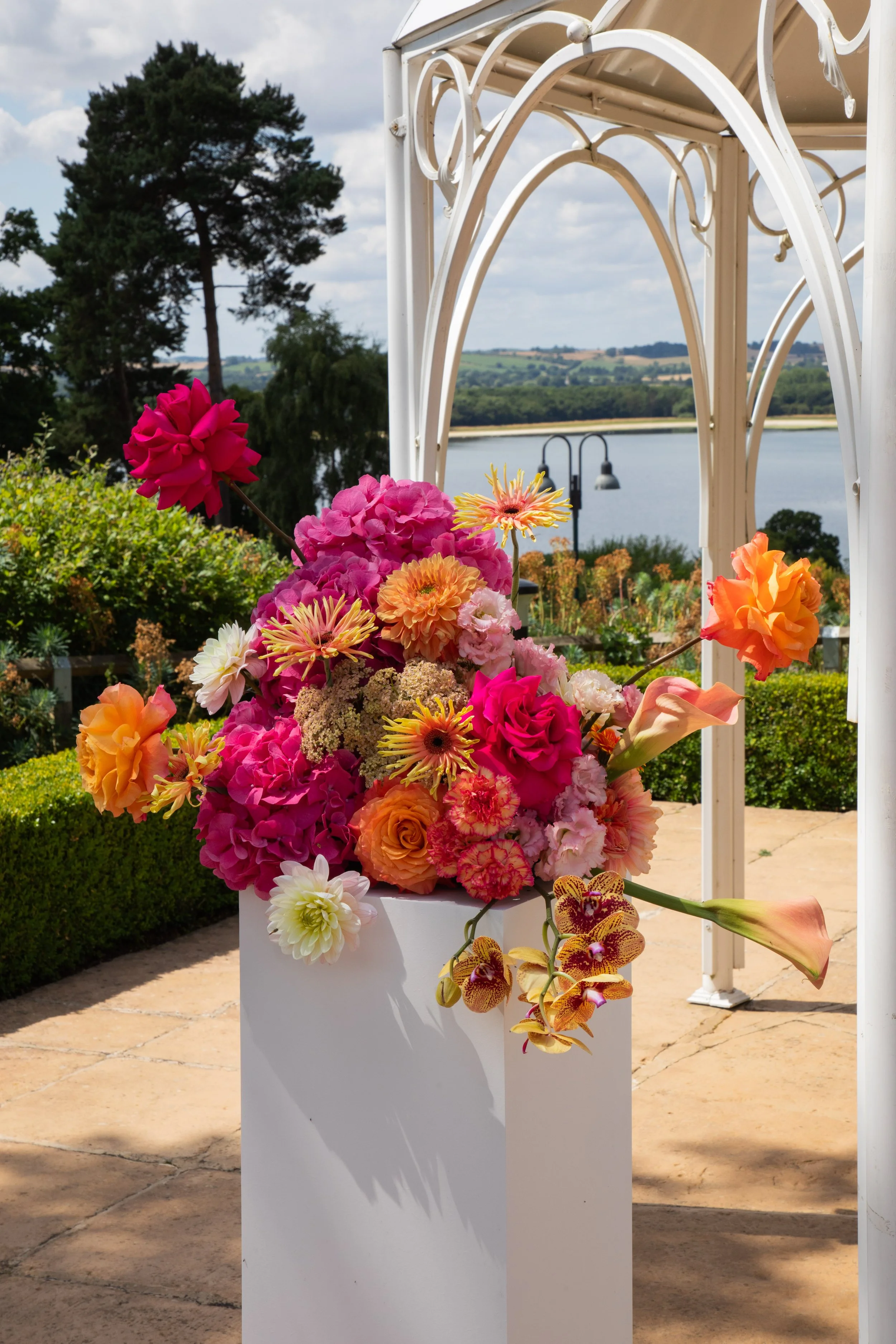Bright and modern floral arrangement by Sweet Pea Sorbet Floristry, featuring vibrant pink, orange, and yellow blooms including roses, orchids, and dahlias. Captured by Harriet Thomas Photography at an outdoor wedding venue overlooking Rutland Water.