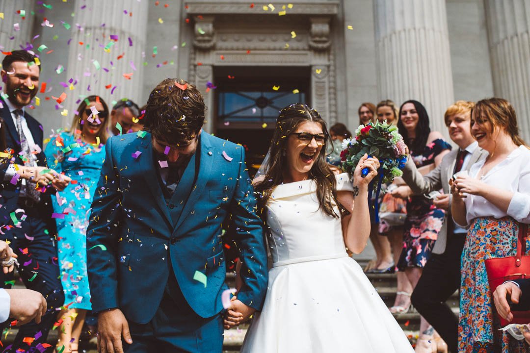 A lively confetti-shot moment captured by Barbara K Photography, showing newlyweds stepping out of a grand London ceremony venue as guests shower them in colourful confetti. The bride, wearing a chic off-the-shoulder dress and glasses, beams with pur