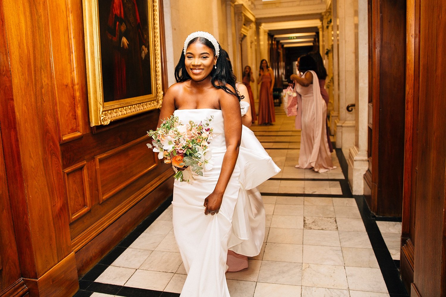 A joyful Black bride walks down an ornate hallway at her London wedding, holding a pastel bouquet of orchids, roses and greenery. Wearing a modern strapless white gown with a statement bow and a pearl headband, she beams at the camera while bridesmai