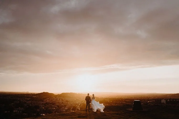 A breathtaking moment high above the city, captured with F.D Young Photography’s cinematic touch. The couple stands together at the edge of the landscape, their silhouettes glowing as the sun spills golden light across the horizon. It’s quiet, expans