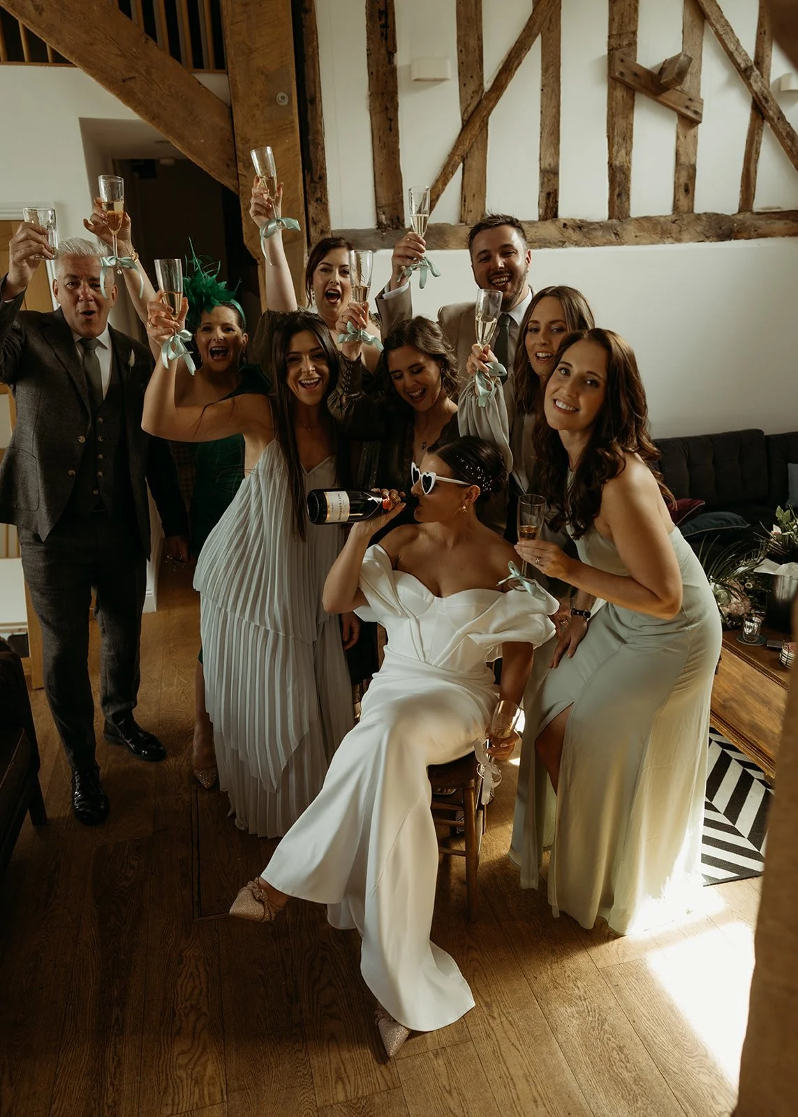 A bride in white sunglasses sits laughing with a champagne bottle in hand while friends and family crowd around her in a rustic room with exposed beams, everyone raising flutes tied with pale ribbons, their pastel and neutral outfits glowing in the w