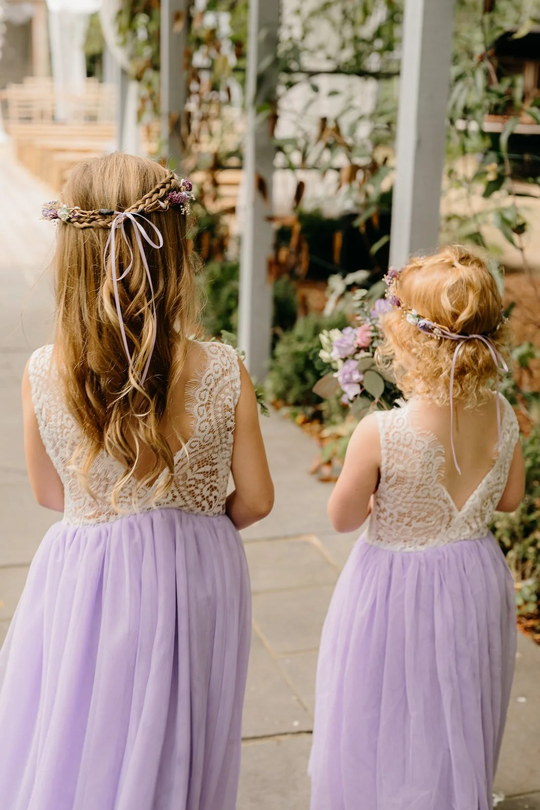 2 little bridesmaids are walking down the aisle with their backs to the camera. They are wearing beautiful dresses with a lilac skirt, and lace back. They are also wearing flower crowns.