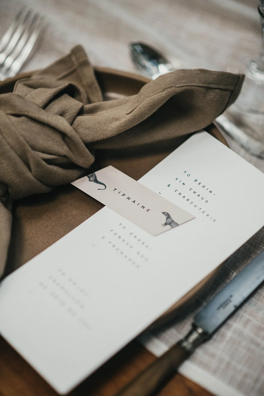 A stylish wedding tablescape detail featuring a modern minimalist menu card paired with a personalised place card illustrated with playful animal motifs placed atop earthy ceramic tableware with a loosely knotted taupe linen napkin creating a refined