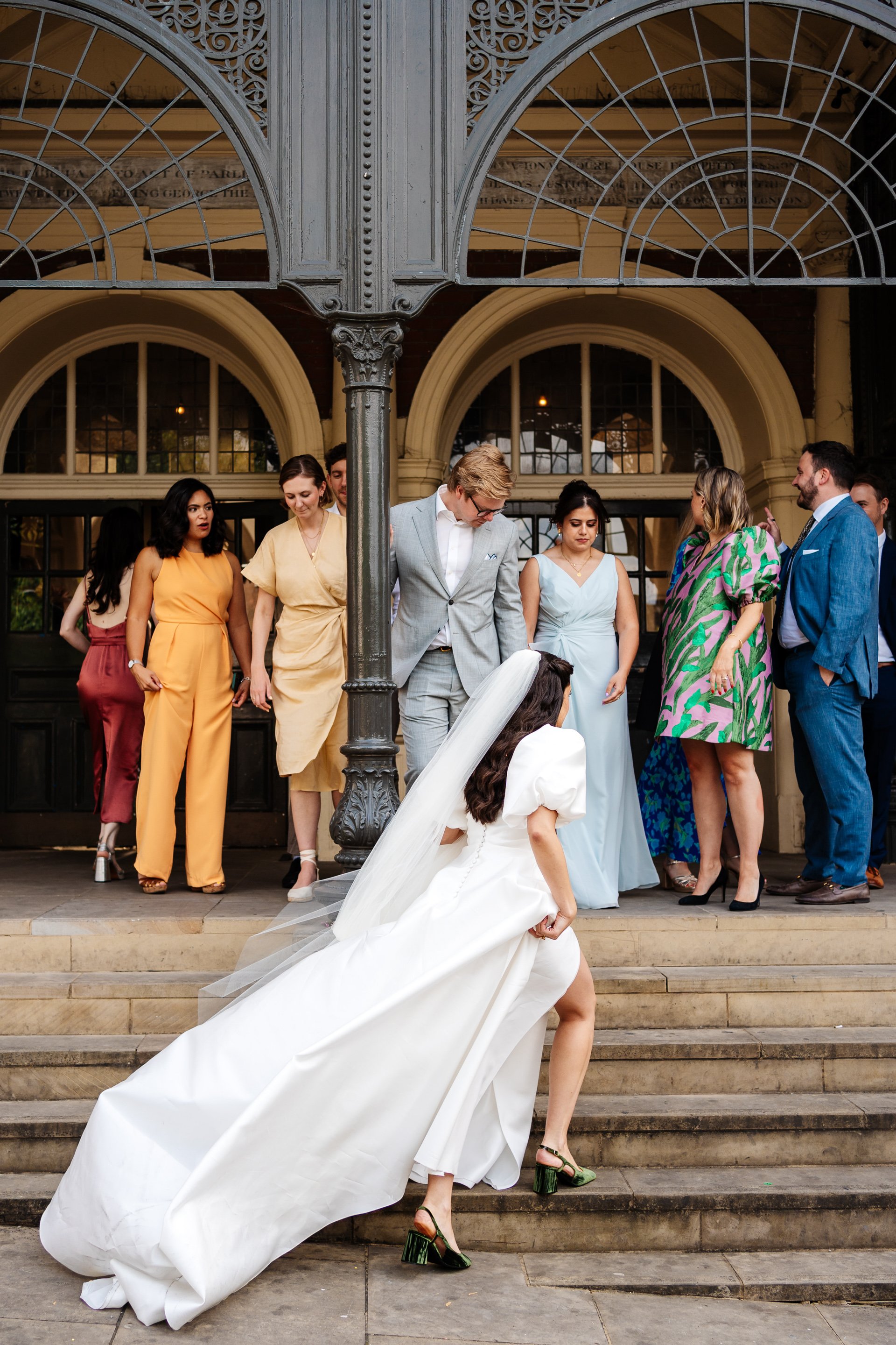 A woman in a long white dress with a flowing train and veil is pictured climbing stone steps outside a historic building with ornate ironwork, while a group of guests in colourful formal outfits stand above her, watching and chatting. The scene shows