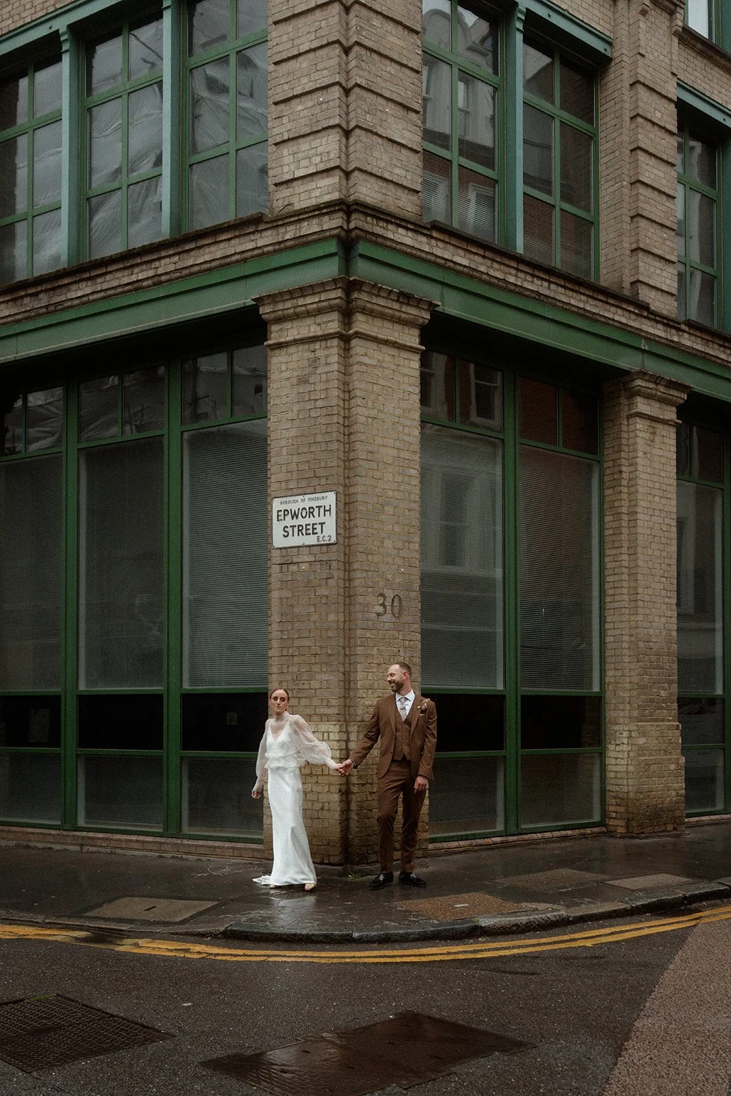 A wedding couple stands on a wet London street corner beneath an Epworth Street sign with the bride in a long white dress and sheer sleeves holding hands with the groom who wears a brown suit, positioned in front of a tall brick building with large g