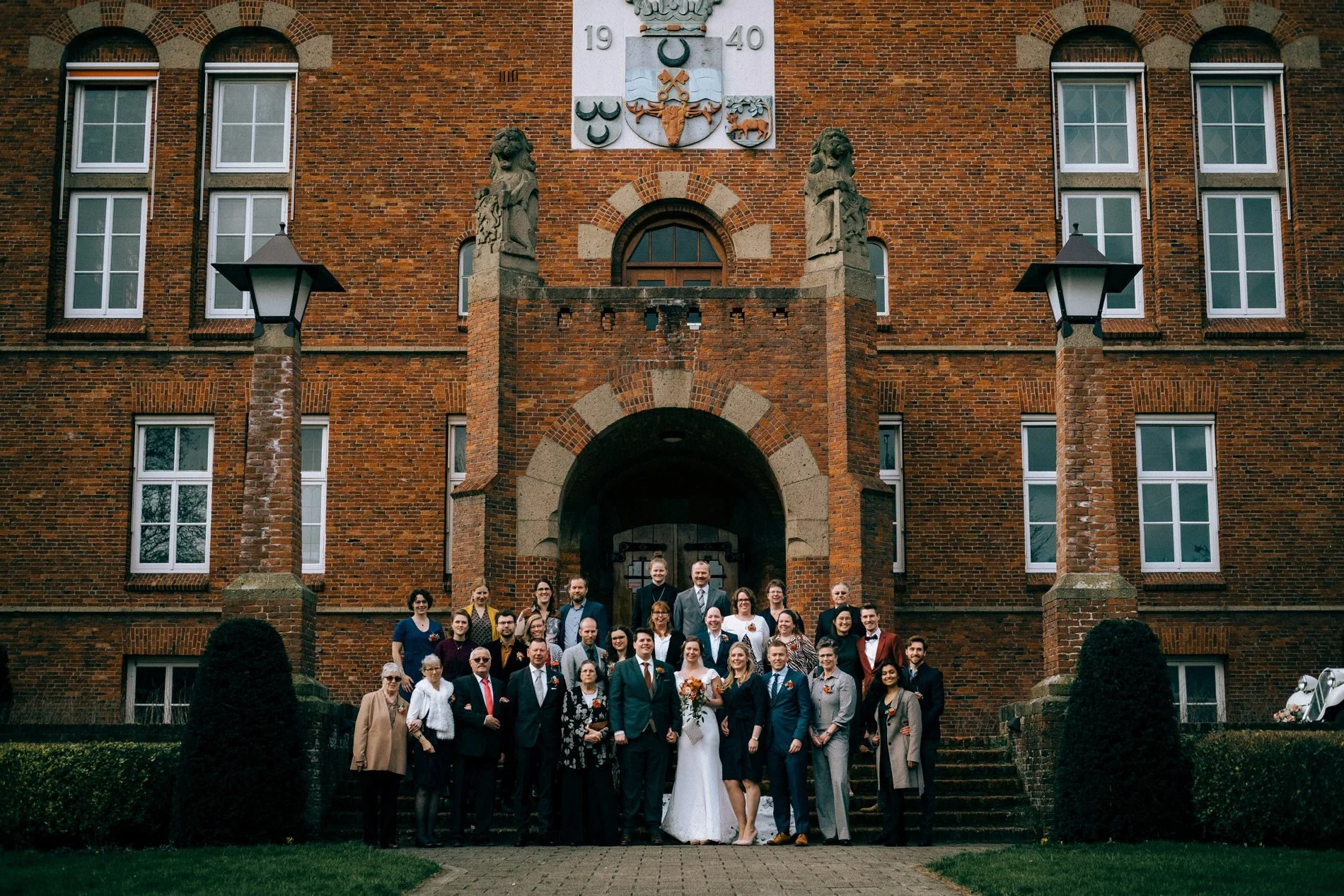 A relaxed group wedding portrait outside a historic brick venue, capturing family and friends in a natural, documentary style by Quiet Mornings Wedding Photography in Scotland.