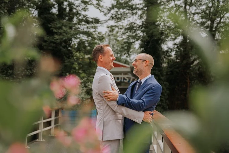 A couple share a warm embrace on a garden walkway surrounded by soft greenery and pink flowers, captured by Joshua Humphrey Photography. Their joyful expressions and natural connection create a relaxed outdoor wedding portrait with a peaceful romanti