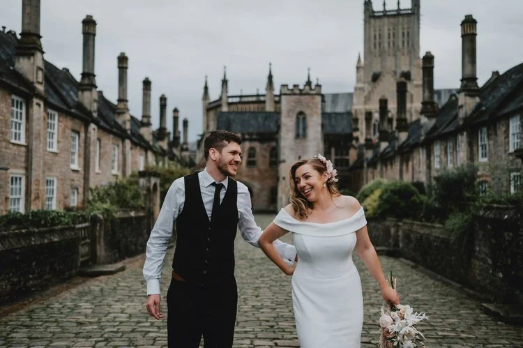 A joyful and romantic wedding portrait captured by Matt Fox Photography, showing a couple walking together along a historic cobblestone street with a grand cathedral rising in the background. The bride wears an elegant off the shoulder gown and carri