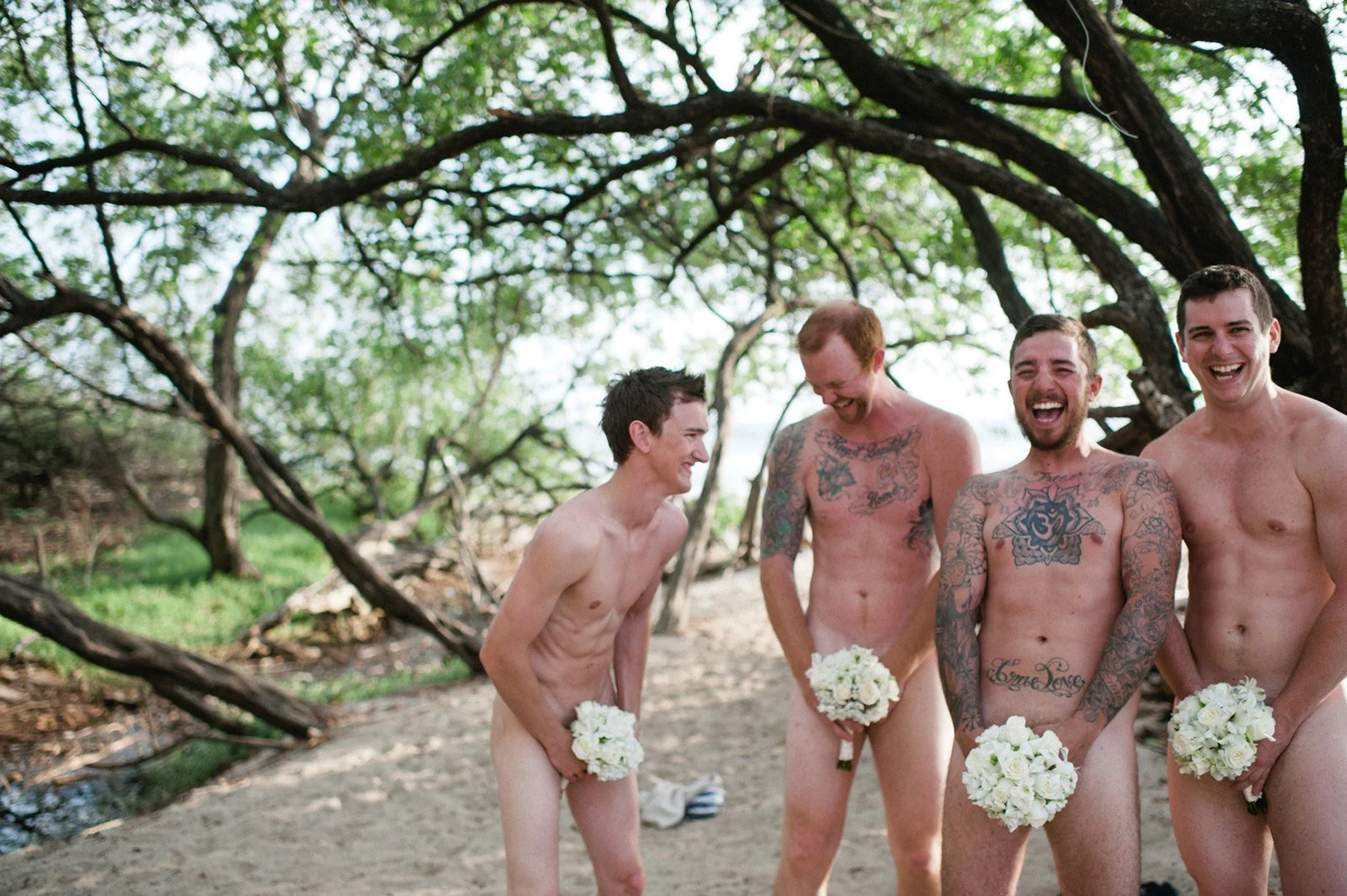 A playful and lighthearted beach moment with groomsmen posing nude, using white bouquets for modesty, laughing together under the trees, captured by The Experiential Wedding.