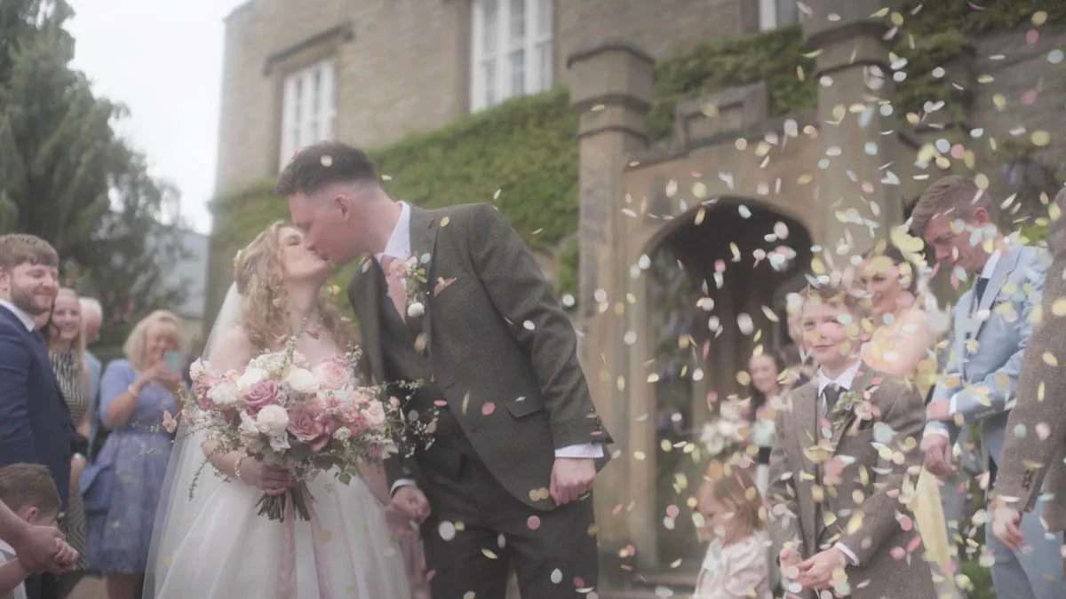 A couple shares a soft kiss beneath a pastel shower of confetti as guests gather around them smiling and celebrating while delicate blooms gentle light and the historic stone backdrop create a dreamy romantic atmosphere.