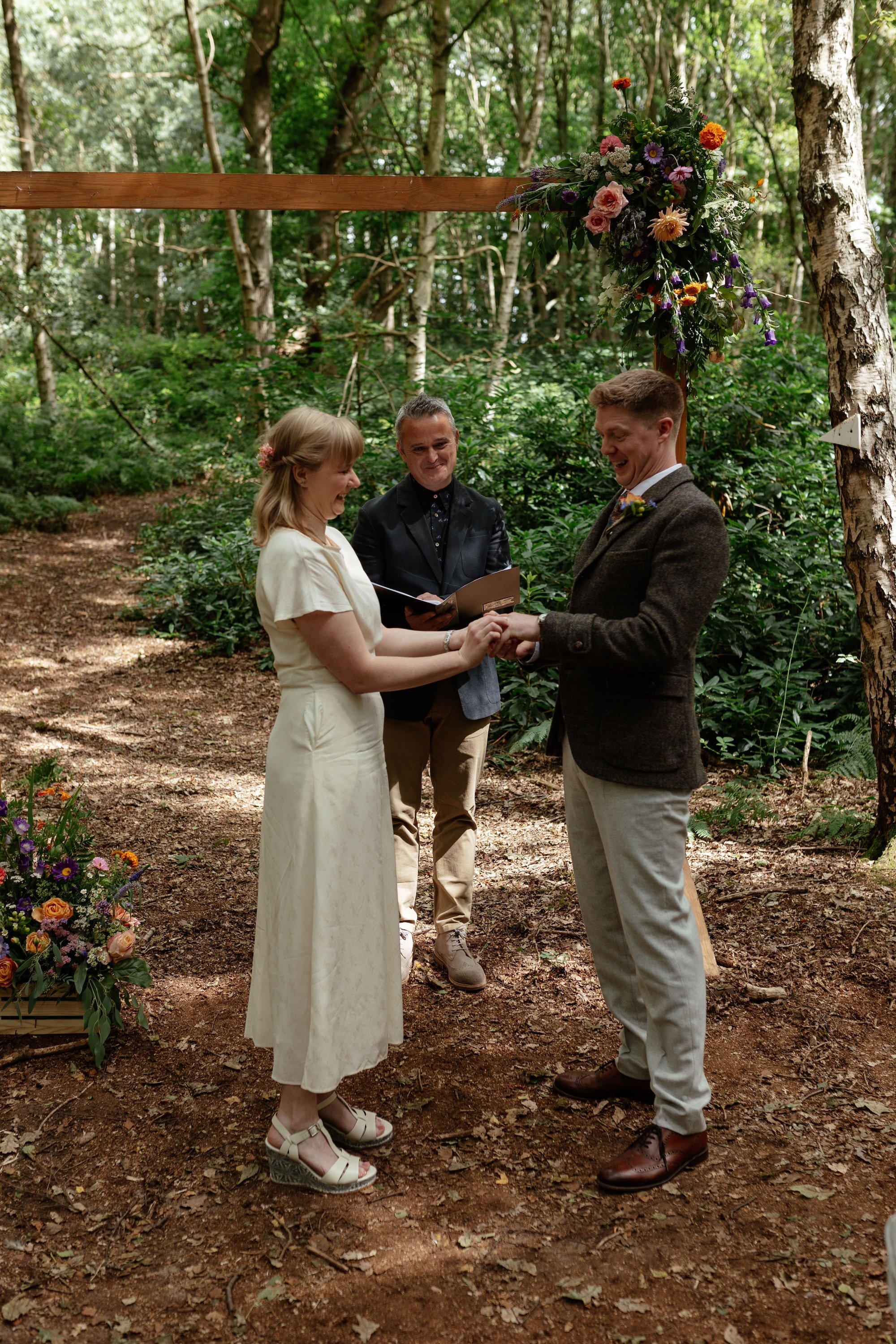 A wooden ceremonial arch with colourful flowers. There is a bride and groom and a male celebrant. They are in the woods.