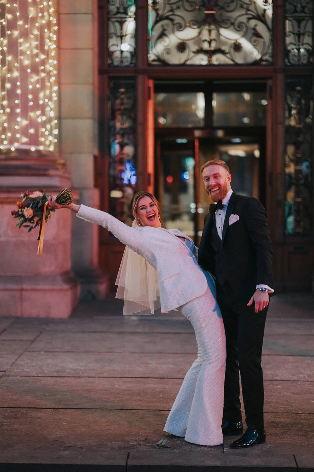 A newlywed couple laugh and celebrate outside an elegant city venue at night, with the bride leaning back playfully in a modern fitted wedding dress and veil while holding her bouquet, captured in a relaxed, documentary wedding photography style by G