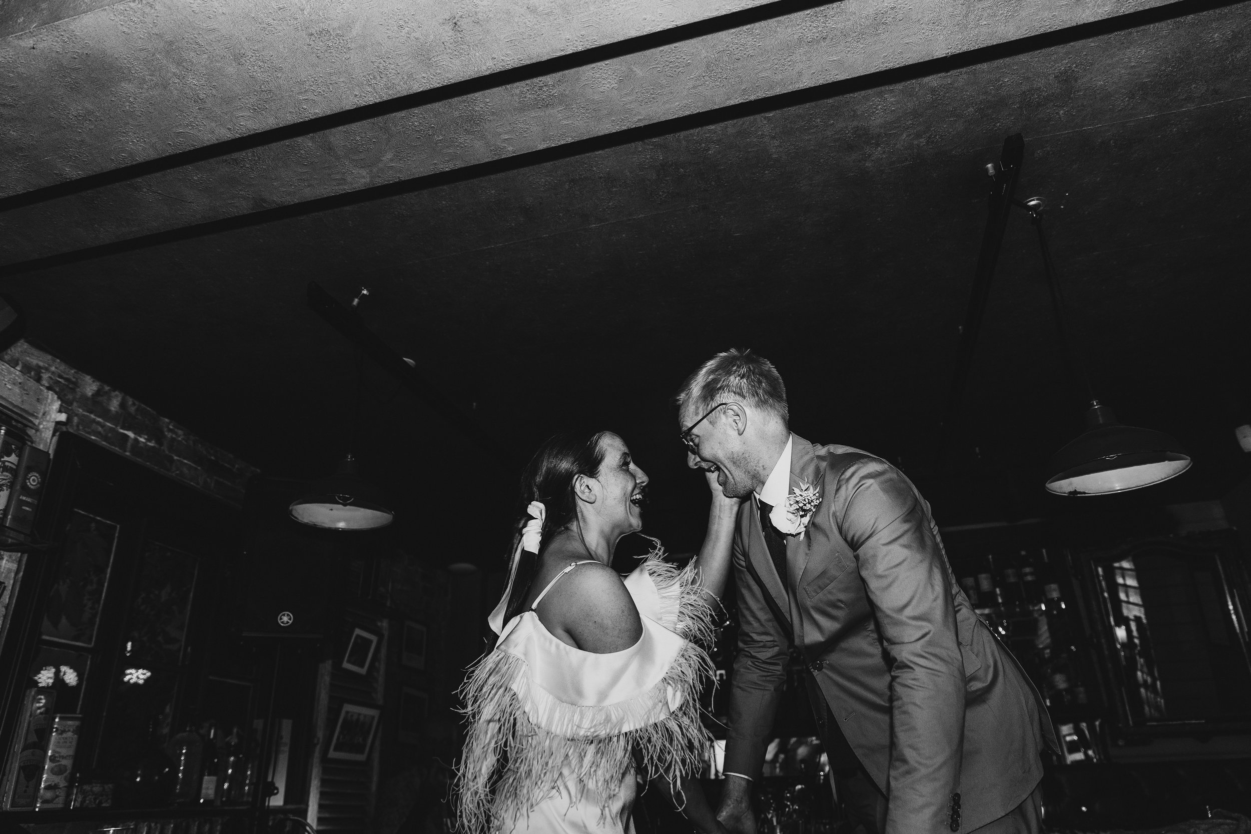 A fun and intimate celebration moment captured by YU Photography, showing the couple laughing together in a dimly lit bar as they lean toward one another with huge smiles. The bride’s feathered dress detail catches the light while she gently holds he