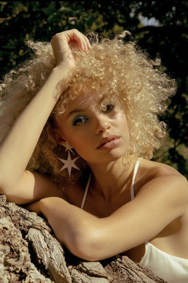 A dreamy outdoor bridal look by Powder Blue Artistry - soft golden skin, natural lips, and ethereal blue eyeshadow accentuated with pearl-like detailing. The voluminous curls and star-shaped earrings add a whimsical, sun-kissed touch, perfect for a m