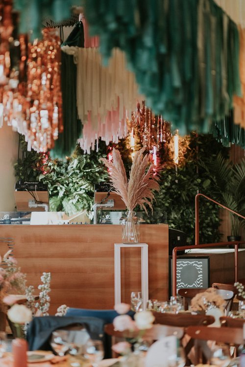 An immersive wedding reception space styled by Streamadelica Event Styling. The design features cascading metallic and fabric fringe garlands in shades of copper, pink, and green, hanging above a modern wooden DJ booth. Lush greenery, pampas grass ar