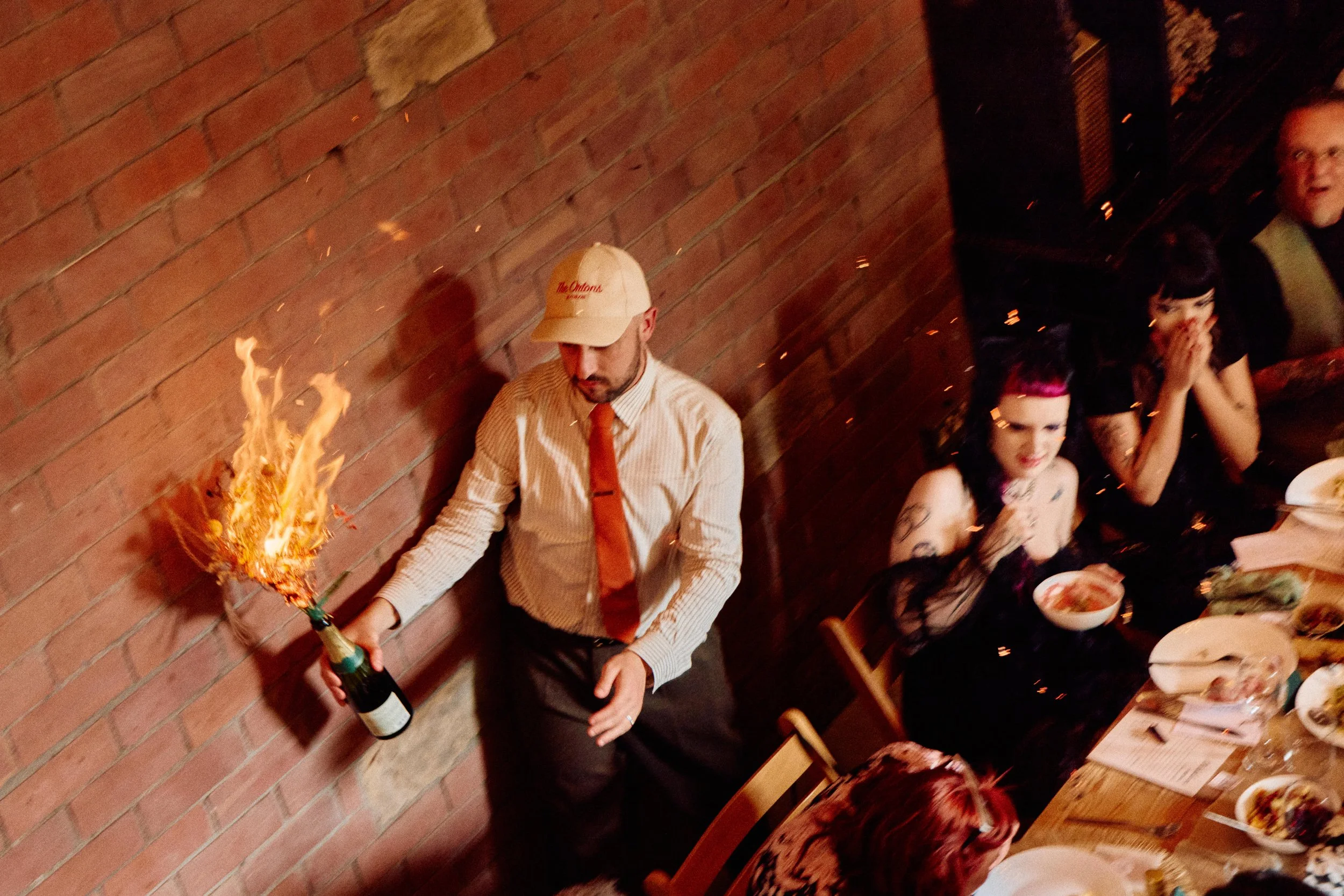 A dramatic Peak District wedding moment as a guest theatrically opens champagne with flames flying, surrounded by cheering diners, captured in bold, candid documentary style by Elizabeth Royse Wedding Photography.