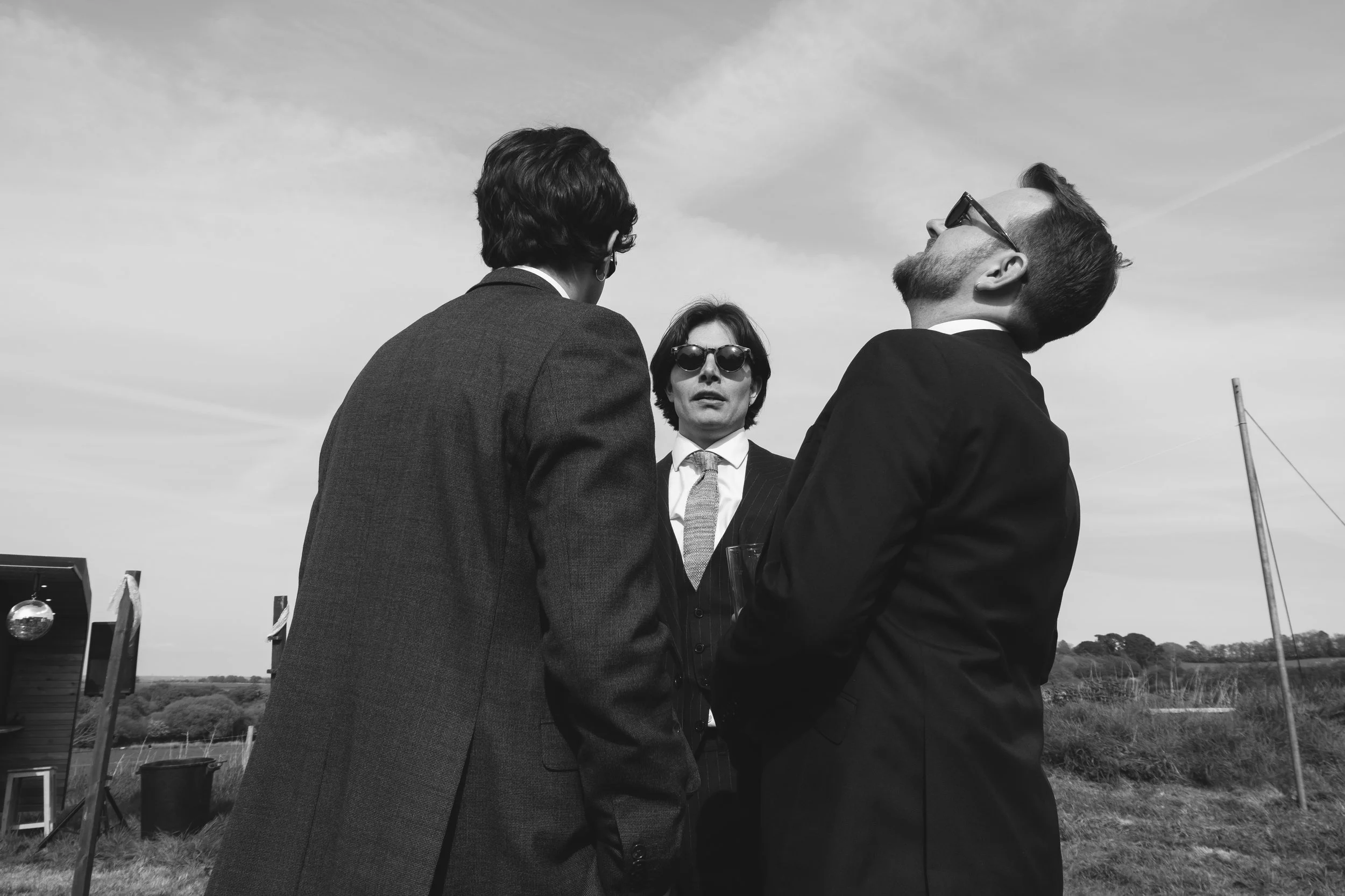 Three wedding guests in suits chat outdoors under a bright sky, captured by Roz Pike Photography. The black and white documentary style and candid expressions create a cool modern moment that reflects the relaxed atmosphere of a contemporary countrys