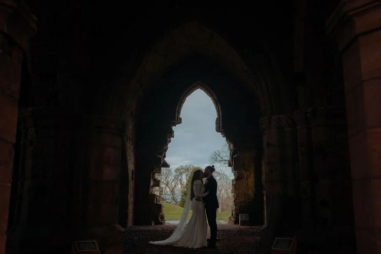 A couple share an intimate kiss beneath the dramatic archway of an ancient stone ruin, silhouetted against soft daylight, beautifully captured by F.D Young Photography. The moody tones, historic architecture and quiet stillness create a powerful, cin