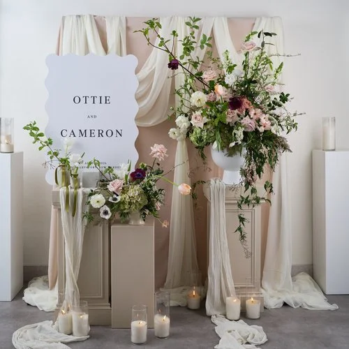Elegant wedding welcome display featuring romantic floral arrangements by Botanique Workshop with blush roses, white anemones, plum accents and natural greenery arranged around a sculptural sign for Ottie and Cameron. Styled with soft draped fabric a