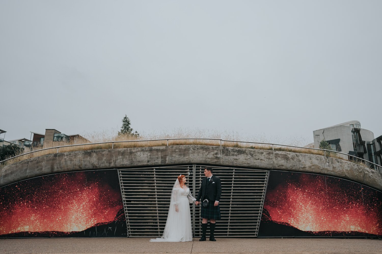 A modern Glasgow wedding portrait by Good Luck Wolf Photography, showing a couple holding hands beneath a concrete bridge, with the bride in a flowing white dress and veil and the groom wearing a traditional Scottish kilt, set against bold urban artw