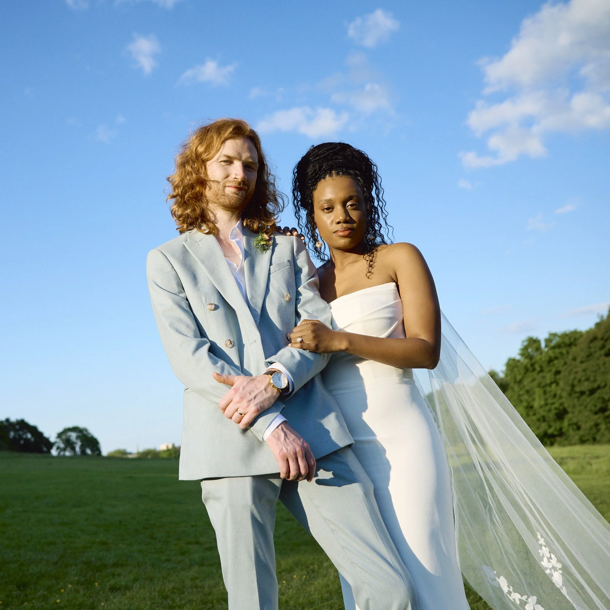 A stylish and modern wedding portrait captured by Wayne La Photography, showing the couple standing close together beneath a clear blue sky in an open field. The groom’s powder blue suit and relaxed pose contrast beautifully with the bride’s elegant 