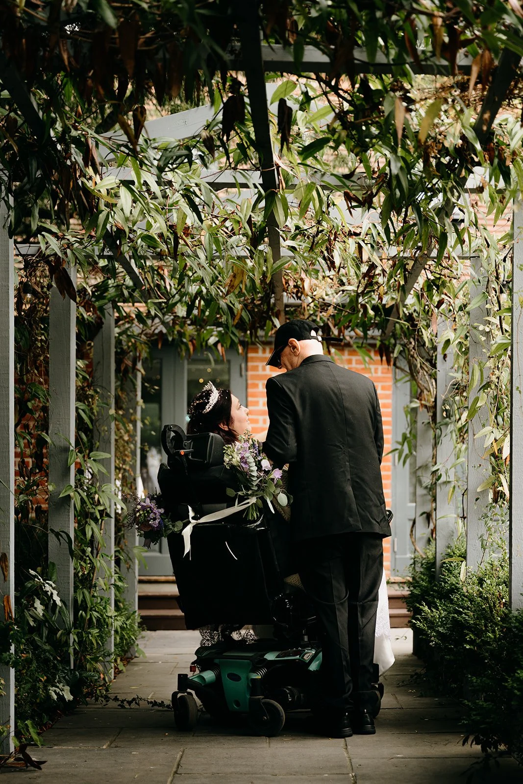 A bride and groom making their way back down the aisle together after their wedding ceremony. Their backs are to the camera and they are surrounded by greenery. The bride's wheelchair has been decorated with flowers.