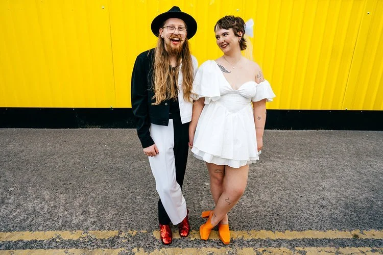 A newly married couple share a relaxed, joyful moment in front of a bright yellow industrial backdrop, captured with vibrant character by Jordanna Marston Photography. Their expressive outfits - from glittering shoes to a playful mini wedding dress -